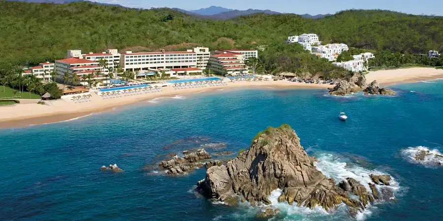 Waves crash into a giant rock outcrop as an all-inclusive beach resort sits in the background along the shore